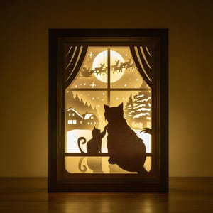 May include: A wooden shadow box with a light inside, depicting a winter scene with two cats looking at each other. The scene includes a snowy landscape, a house, and a flying Father Christmas with reindeer.
