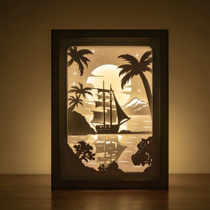 Könnte beinhalten: Ein weißes Papier-Schnittbild in einem Holzrahmen mit beleuchtetem Hintergrund. Die Kunst zeigt ein Segelboot, das auf einem ruhigen Meer mit Palmen und einem Vollmond im Hintergrund segelt.