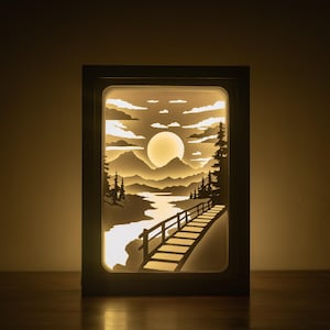 Könnte beinhalten: Ein gerahmtes, beleuchtetes Papierkunstwerk, das eine ruhige Landschaft darstellt. Das Kunstwerk zeigt eine Brücke, Berge, Bäume und einen großen Mond, alles in Weiß- und Beigetönen. Das Stück wird von innen beleuchtet und erzeugt ein warmes, einladendes Leuchten.