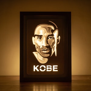 May include: A black framed light-up portrait of Kobe Bryant with his name in white letters below the image.