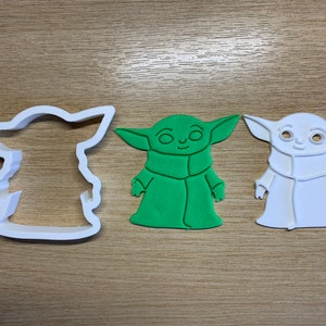 May include: A white plastic cookie cutter and two cookies shaped like a character with large ears and a robe. One cookie is green, and the other is white. The items are on a light brown wooden surface.