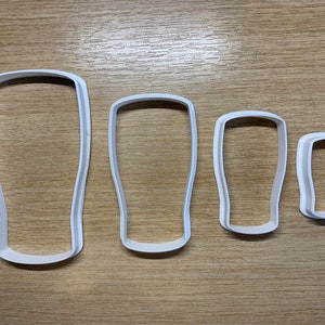May include: Four white plastic cookie cutters shaped like pint glasses. The cutters are arranged by size, from large to small, with a simple design. They are on a light brown wooden surface.
