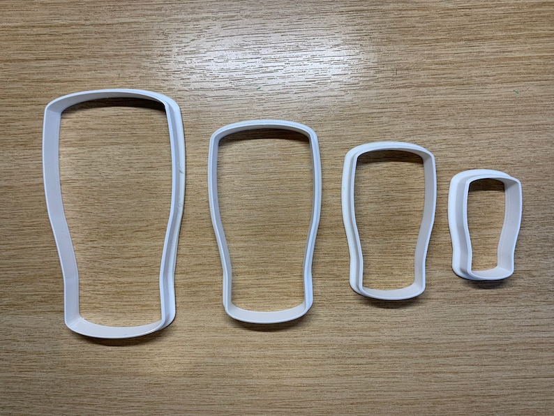 May include: Four white plastic cookie cutters shaped like pint glasses. The cutters are arranged by size, from large to small, with a simple design. They are on a light brown wooden surface.