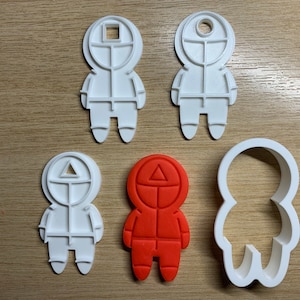 May include: A collection of white and red plastic cookie cutters in the shape of figures with geometric shapes on their heads. The cutters include a square, circle, and triangle. One cutter is red, and the others are white.