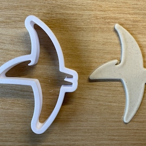 Peut inclure: Un emporte-pièce blanc en forme d'oiseau en plastique à côté d'un biscuit en forme d'oiseau. Le découpoir a un contour détaillé des ailes et de la queue de l'oiseau. Le biscuit est brun clair et de forme similaire.