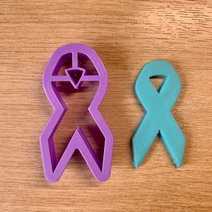 Op de afbeelding: Een paarse lintvormige koekjesuitsteker naast een turquoise lintvormig koekje. De koekjesuitsteker is van plastic en heeft een geometrisch ontwerp. De turquoise koek is gemaakt van deeg.