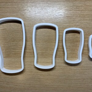 May include: Four white plastic cookie cutters shaped like pint glasses. The cutters are arranged by size, from large to small, with a simple, rounded design. They are on a light brown wooden surface.