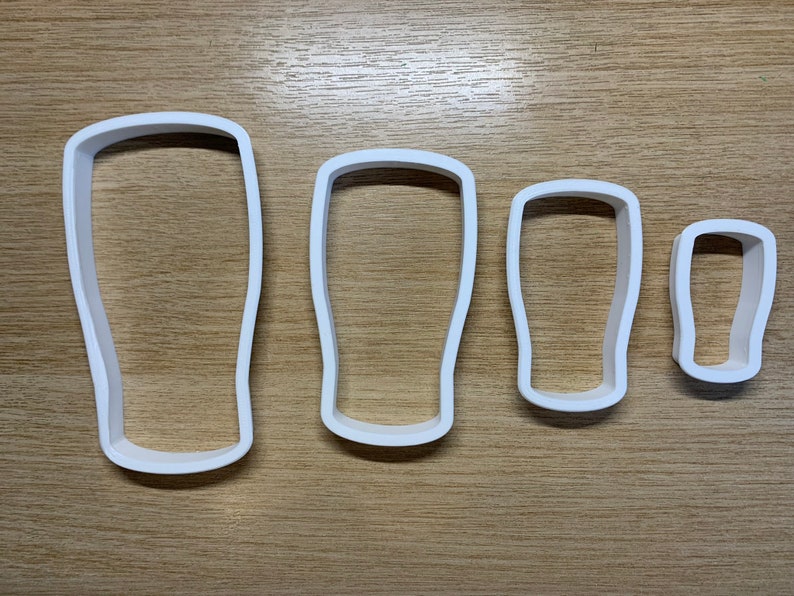 May include: Four white plastic cookie cutters shaped like pint glasses. The cutters are arranged by size, from large to small, with a simple, rounded design. They are on a light brown wooden surface.