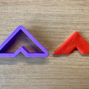 May include: A purple plastic cookie cutter in the shape of an angled arrow is next to a small, red, angled arrow made of a food product. Both are on a light brown wooden surface. The cookie cutter has a triangular shape with a notch cut out of the bottom.