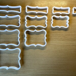 May include: A selection of white plastic cookie cutters in different sizes and shapes. The cutters feature a rectangular design with decorative edges, ideal for baking. The cutters are arranged on a light brown wooden surface.
