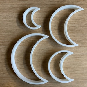 Puede incluir: Cuatro cortadores de galletas blancos en forma de media luna de varios tamaños están dispuestos sobre una superficie de madera de color marrón claro. Los cortadores están hechos de un material liso y sólido y están diseñados para hornear o manualidades.