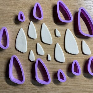 Puede incluir: Un surtido de cortadores de pendientes de arcilla polimérica y piezas de arcilla blanca dispuestas sobre una superficie de madera. Los cortadores son de color lila claro y tienen varias formas y tamaños. Las piezas de arcilla tienen forma de lágrima.