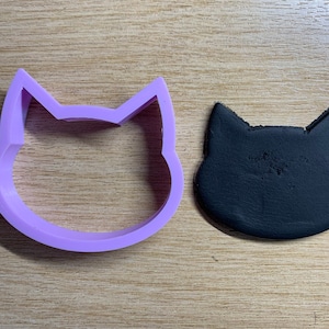 May include: A light purple cat-shaped cookie cutter next to a black cat-shaped cookie. The cookie cutter is made of plastic and has a simple outline of a cat's head. The black cookie is made of dough.