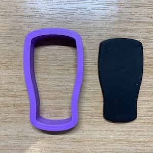 May include: A purple plastic cookie cutter in the shape of a pint glass sits next to a black foam insert, both on a wooden surface. The cookie cutter has a rectangular shape with rounded edges.