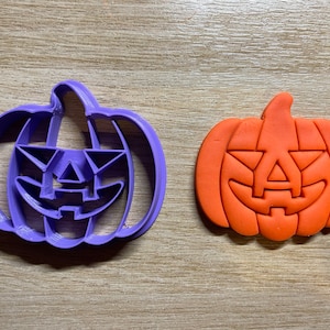 May include: A purple plastic pumpkin-shaped cookie cutter next to an orange pumpkin-shaped cookie. The cookie cutter has a jack-o'-lantern face design. The orange cookie has the same jack-o'-lantern face design.