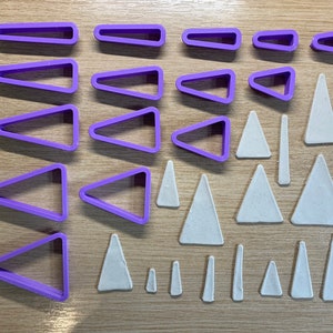 Pode incluir: Uma vista aérea mostra uma coleção de cortadores de biscoitos geométricos de plástico roxo e formas de argila bege. Os cortadores têm várias formas, incluindo triângulos e retângulos. As formas de argila também são triângulos e outras formas geométricas.