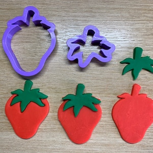 Könnte beinhalten: Ein Set aus Erdbeer-Ausstechern und Tonmodellen. Die Ausstecher sind lila, und die Ton-Erdbeeren sind rot mit grünen Oberteilen. Das Set enthält eine Erdbeerkontur, ein Blatt und eine Beerenoberseite.