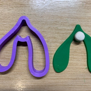 May include: A purple plastic mistletoe cookie cutter next to a green mistletoe shape with a white ball. The cookie cutter is a single piece with a curved design. The green mistletoe shape has a white ball in the centre.