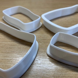 May include: Four white plastic cookie cutters with a geometric shape. The cutters are arranged on a light brown wooden surface. The cutters have a smooth, curved design, and are likely used for baking or crafting.