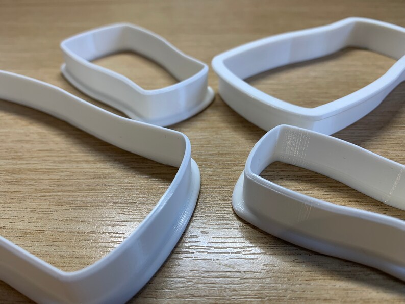 May include: Four white plastic cookie cutters with a geometric shape. The cutters are arranged on a light brown wooden surface. The cutters have a smooth, curved design, and are likely used for baking or crafting.