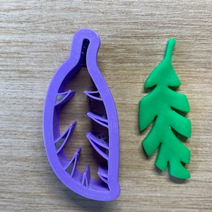 Op de afbeelding: Een paarse blad-vormige koekjesvorm naast een groene blad-vormige koekje. De koekjesvorm is gemaakt van plastic en heeft een gedetailleerd ontwerp. De groene koekje is effen van kleur en heeft een gedetailleerde bladvorm.