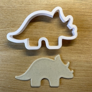 Peut inclure: Un emporte-pièce en plastique blanc en forme de dinosaure, à côté d'un biscuit beige découpé dans la même forme. Le découpoir est un contour détaillé d'un triceratops, un herbivore préhistorique à trois cornes.