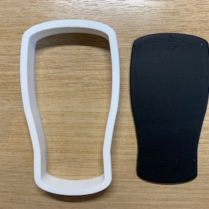 May include: A white plastic cookie cutter in the shape of a pint glass sits next to a black, textured piece of material, also in the shape of a pint glass. The items are on a light brown wooden surface.