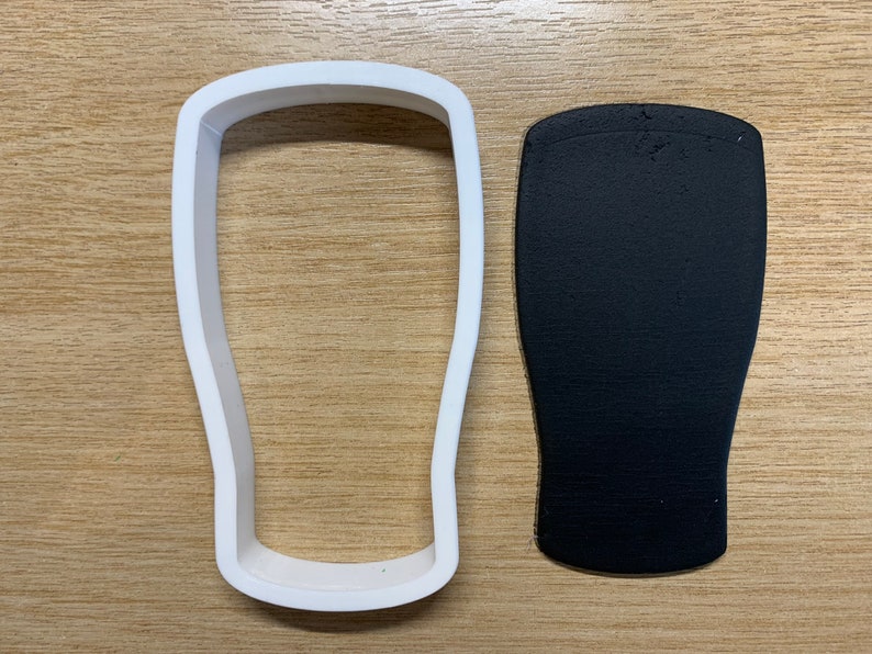 May include: A white plastic cookie cutter shaped like a pint glass is next to a black, textured piece of material, also in the shape of a pint glass. The items are on a light brown wooden surface.