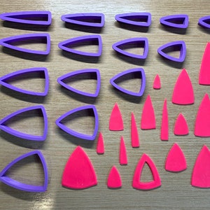 Può includere: Una collezione di formine per biscotti a forma di triangolo viola e taglierine per argilla a forma di triangolo rosa sono disposte su una superficie di legno chiaro. Le taglierine variano in dimensioni e forma, adatte per l'artigianato o la cottura.