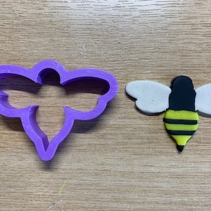May include: A purple plastic bee-shaped cookie cutter next to a handmade bee. The bee has white wings, a black head and stripes of yellow and black on its body. The items are on a light brown wooden surface.