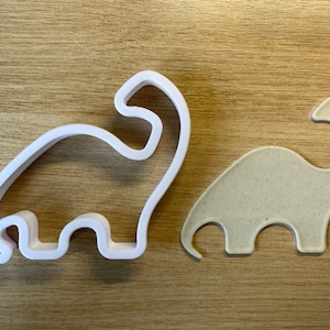 Peut inclure: Un emporte-pièce en plastique blanc en forme de dinosaure à côté d'une découpe en forme de dinosaure beige. L'emporte-pièce a un contour détaillé du corps du dinosaure, y compris sa tête, son long cou et ses pattes. La découpe est une forme pleine du même dinosaure.