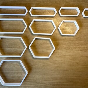 Puede incluir: Un conjunto de cortadores de galletas geométricos blancos de varios tamaños y formas, incluyendo rectángulos y hexágonos. Los cortadores están dispuestos sobre una superficie de madera marrón claro.