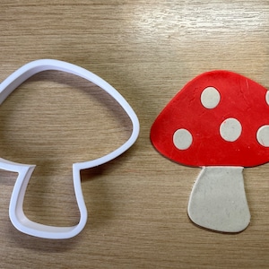 Puede incluir: Un cortador de galletas blanco con forma de hongo junto a un hongo rojo con lunares blancos. El hongo está hecho de arcilla o fondant. El fondo es una superficie de madera marrón claro.