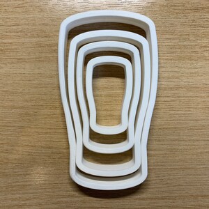 May include: A white plastic cookie cutter set in the shape of a pint glass. The set includes four nested cutters, each slightly smaller than the last. The cutters are arranged on a light brown wooden surface.