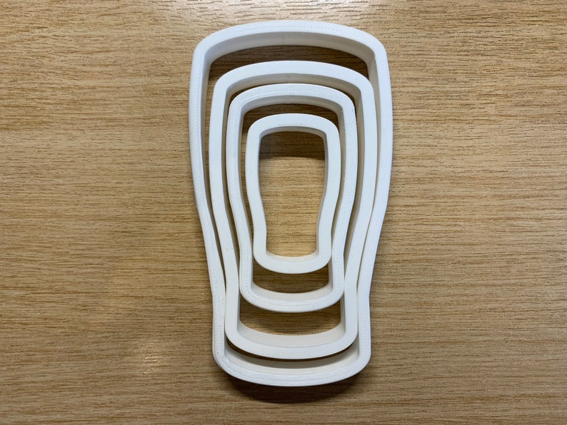 May include: A white plastic cookie cutter set in the shape of a pint glass. The set includes four nested cutters, each slightly smaller than the last. The cutters are arranged on a light brown wooden surface.