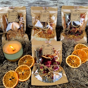 May include: A wicker basket filled with clear bags of dried botanicals, a lit candle, and dried orange slices. The bags are labeled "Calming", "Autumn Harvest", "Moonlight Ritual", and "Renewal". The candle is in a teal ceramic holder.
