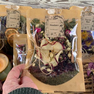 May include: Four clear plastic bags filled with dried herbs, flowers, and fruit. The bags are tied with twine and labeled with handwritten tags. The tags read "Cleansing", "Abundance", "Peaceful Mind", and "Simmer Pot".