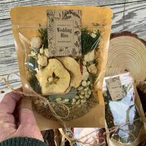 May include: A brown paper bag with a clear plastic window, containing a simmer pot blend. The bag is tied with twine and has a label that reads "Wedding Bliss" with a floral design. The blend includes dried apple slices, flowers, and herbs.