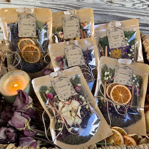 May include: A wicker basket filled with six clear plastic bags containing dried herbs, flowers, and fruit. Each bag has a small paper tag with a handwritten title, such as "Grounding" and "Peaceful Mind".