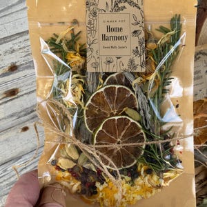 May include: A brown paper bag with a clear plastic window, containing a simmer pot blend. The bag is tied with twine and has a label that reads "Simmer Pot Home Harmony Sweet Molly Jayne's". The blend includes dried citrus, herbs, and spices.