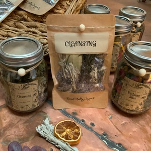 May include: A collection of glass jars filled with dried herbs and flowers, labeled with the names "Cleansing" and "Wedding Bliss". The jars are sitting on a wooden table with a brown paper bag labeled "Cleansing" and a piece of dried sage.