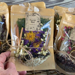 May include: Three brown paper bags with clear plastic windows filled with dried herbs, flowers, and spices. The bags are tied with twine and labeled with the text "Simmer Pot Peaceful Mind" and "Simmer Pot Blessings".