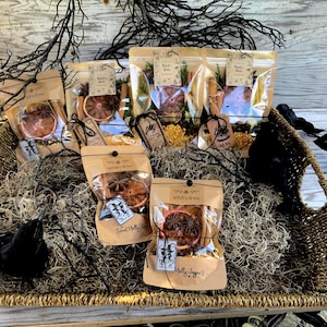 May include: A wicker basket filled with dried herbs, spices, and decorative elements. Several clear bags contain ingredients like orange slices, star anise, and cinnamon sticks. The bags are labeled "Witch's Brew" and are tied with twine. Black decorative branches and a black crow are also in the basket.