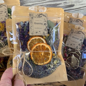 May include: Three clear plastic bags filled with dried herbs, flowers, and fruit slices. The bags are tied with twine and labeled with handwritten tags. The tags read "Peaceful Mind", "Cleansing", and "Blessings".