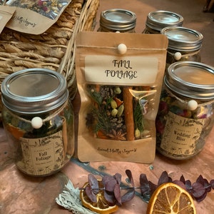 May include: A collection of glass jars filled with dried herbs and spices, labeled with names like "Fall Foliage" and "Wedding Bliss". The jars are arranged on a copper-colored surface with dried orange slices and eucalyptus leaves.
