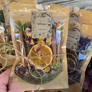 May include: A close-up of a clear plastic bag filled with dried herbs, flowers, and spices. The bag is tied with twine and has a label that reads "Self Love" with a floral design.