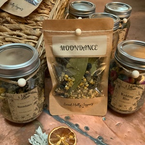 May include: A collection of glass jars filled with dried herbs and flowers. The jars are labeled with names like "Moondance" and "Wedding Bliss". The jars are sitting on a copper-colored surface.