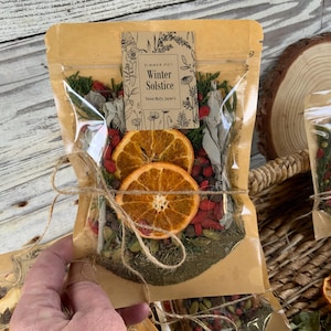 May include: A clear cellophane bag filled with dried herbs, spices, and orange slices, tied with twine. The bag has a brown paper label that reads "Winter Solstice". The image is set against a rustic wooden background.