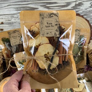 May include: A brown paper bag with a clear window, tied with twine, contains dried apple slices, cinnamon sticks, star anise, and vanilla beans. The bag has a label that reads "Summer Pot Warm Apple Pie".