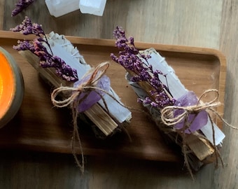 May include: Two bundles of sage smudge sticks tied with twine and decorated with dried lavender and amethyst crystals. The bundles are on a wooden tray.